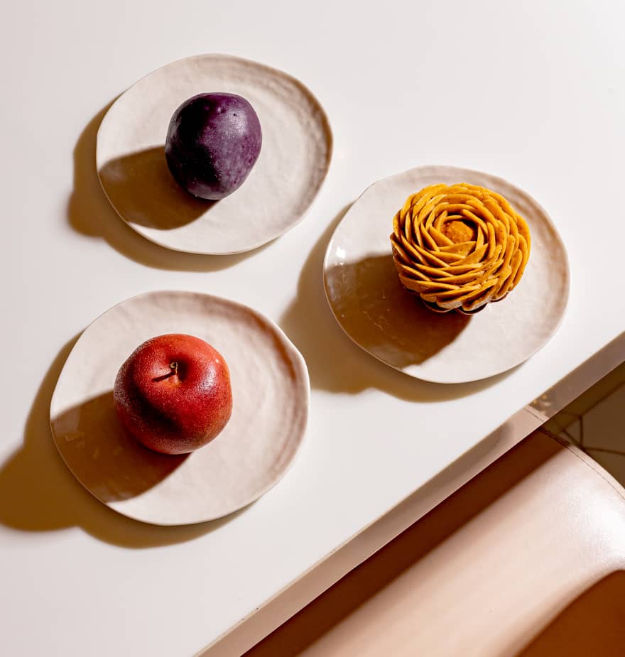 Three plated fruit-inspired pastries, shaped like a plum, apple, and spiraled tart, arranged on a glossy white table.