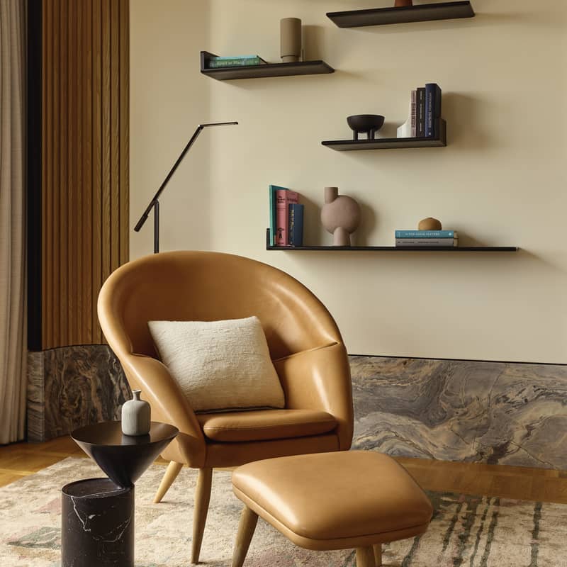 Stylish lounge corner with tan leather armchair, footstool, side table, and minimalist floating shelves on a neutral wall.