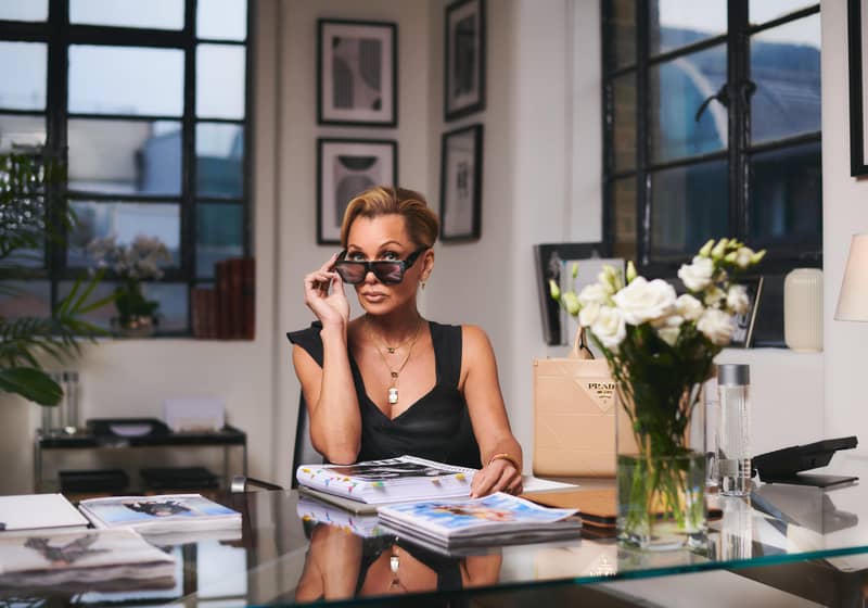 A poised woman seated at a stylish desk, glancing over her sunglasses with a confident expression. Surrounded by magazines, flowers, and designer accessories, she exudes sophistication and authority.