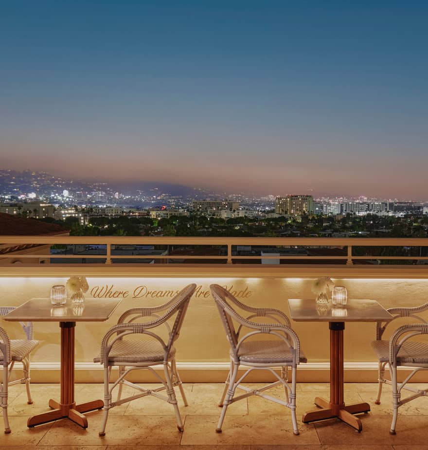 Rooftop terrace with bistro tables, city skyline glowing at dusk, and “Where Dreams Are Made” script on the wall.