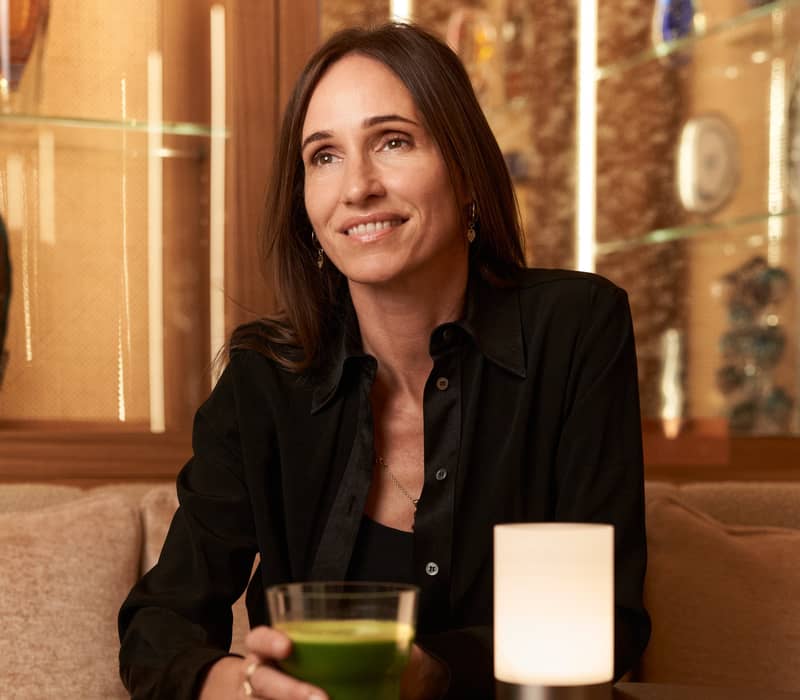 A woman sits in an elegant lounge, holding a green juice, surrounded by warm lighting, plush seating, and decorative glass accents.