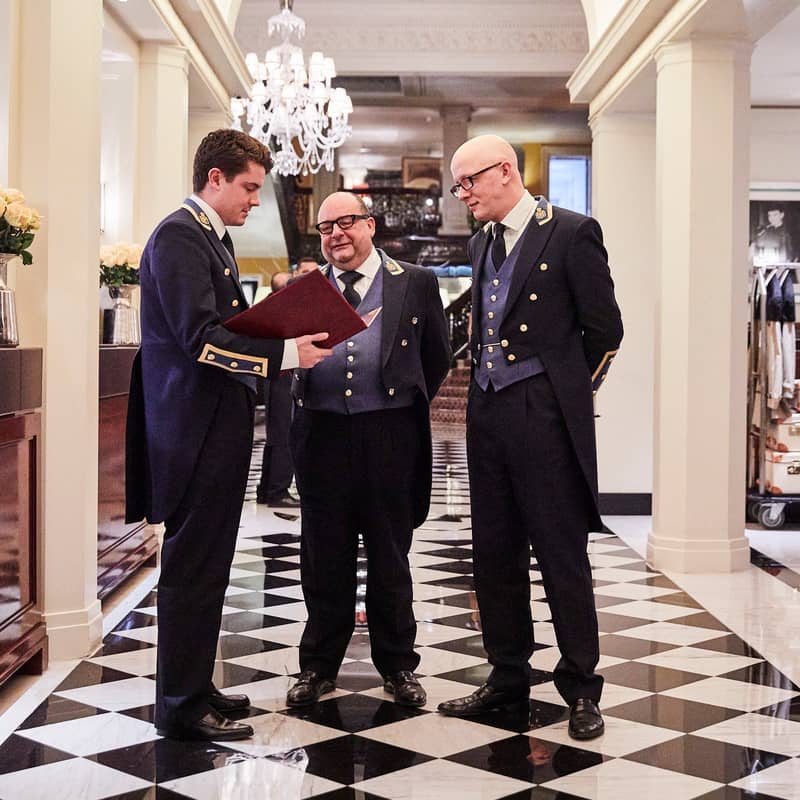 Three concierges in distinguished uniforms discuss guest arrangements in a grand hotel lobby with checkered flooring and chandeliers.