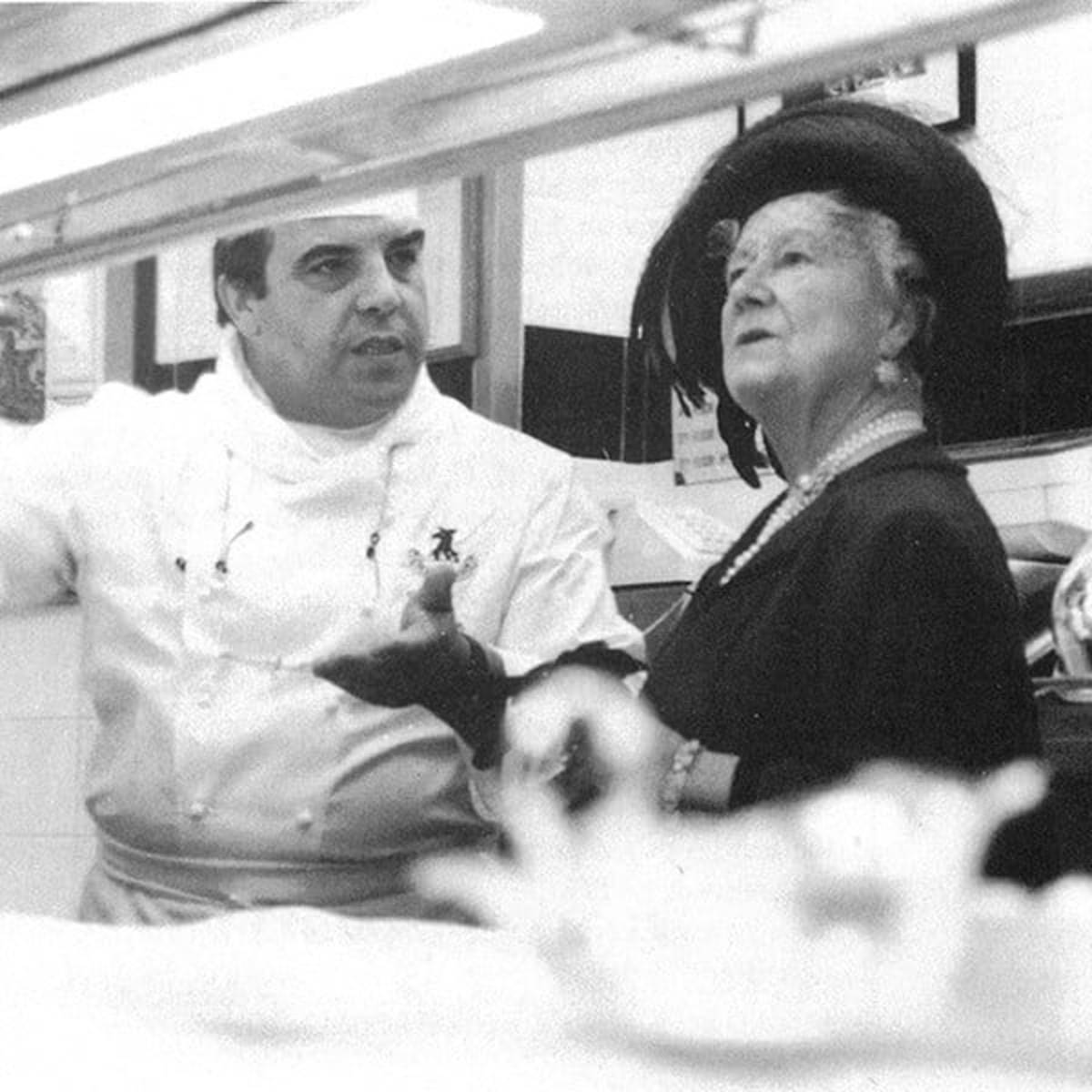 Vintage black-and-white photo of a chef conversing with an elegantly dressed lady in a grand hat, exuding timeless charm
