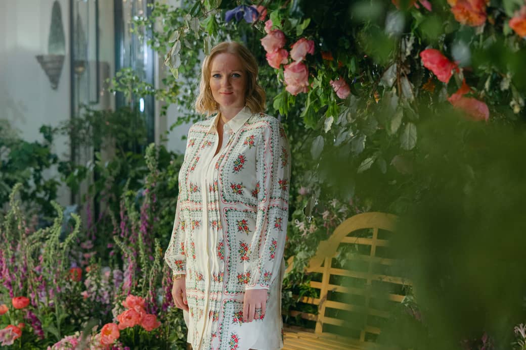 A woman stands gracefully in a floral-themed setting, surrounded by lush greenery and vibrant blossoms, exuding elegance in a patterned dress and espadrille wedges.