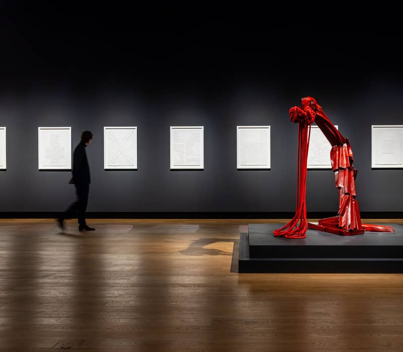 Person walking through a modern gallery with white framed artworks and a striking red abstract sculpture on display.