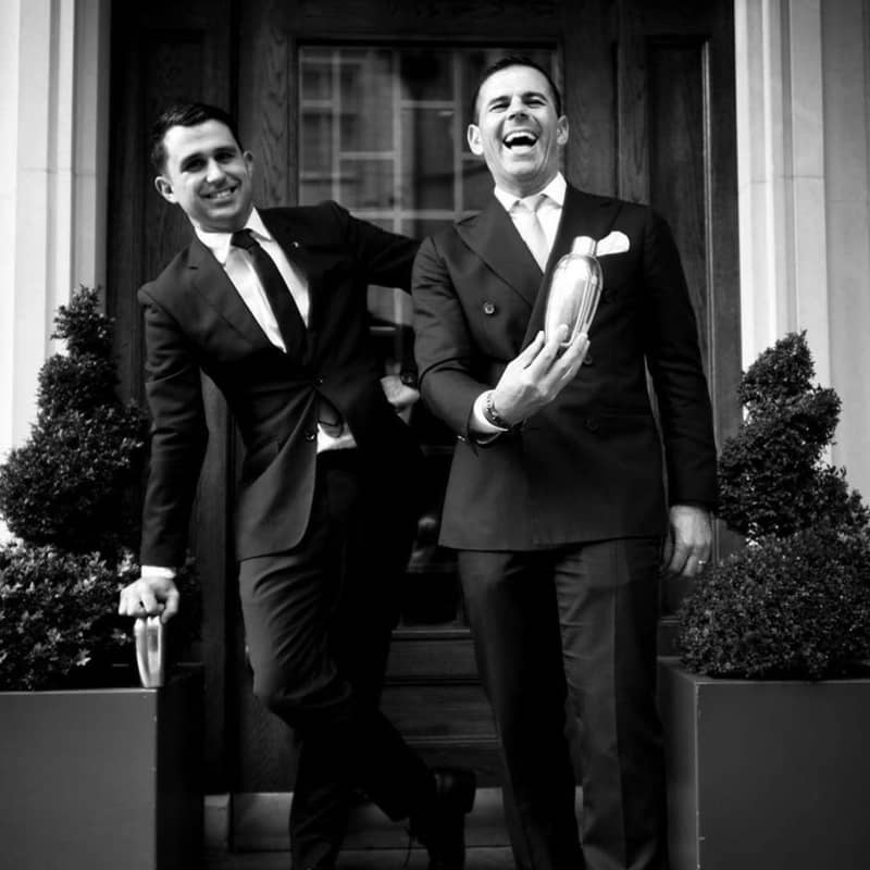 Two bartenders in tailored suits laugh outside, holding cocktail shakers, exuding charm and hospitality.