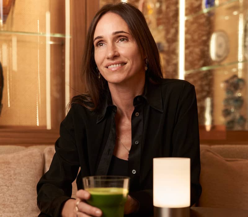 A woman sits in an elegant lounge, holding a green juice, surrounded by warm lighting, plush seating, and decorative glass accents.