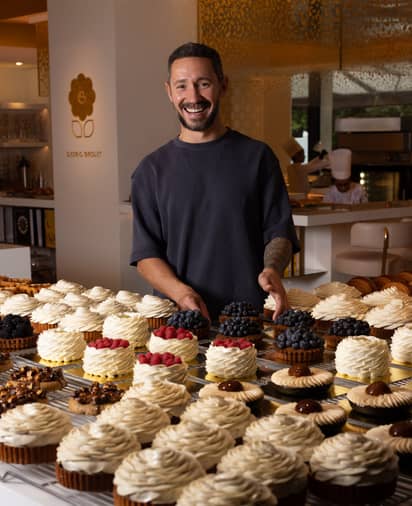 Pastry chef Cédric Grolet smiles behind a display of exquisite tarts, adorned with whipped cream swirls and fresh berries.