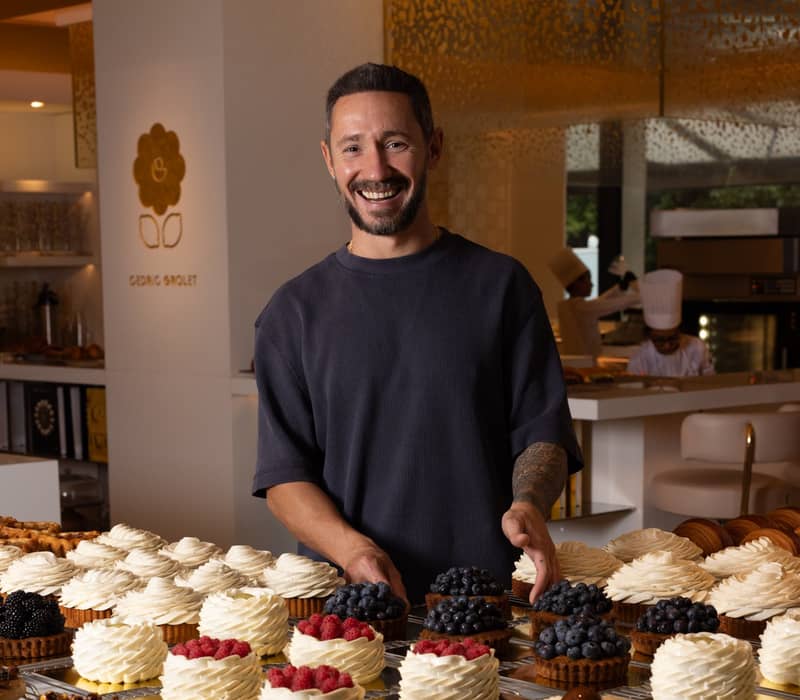 Pastry chef Cédric Grolet smiles behind a display of exquisite tarts, adorned with whipped cream swirls and fresh berries.