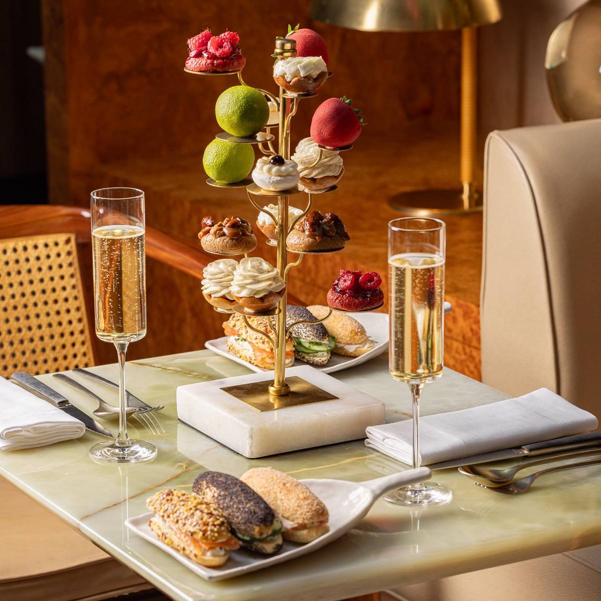 Afternoon tea setup with pastries on a tiered stand, savoury bites, and two glasses of champagne on a marble table.