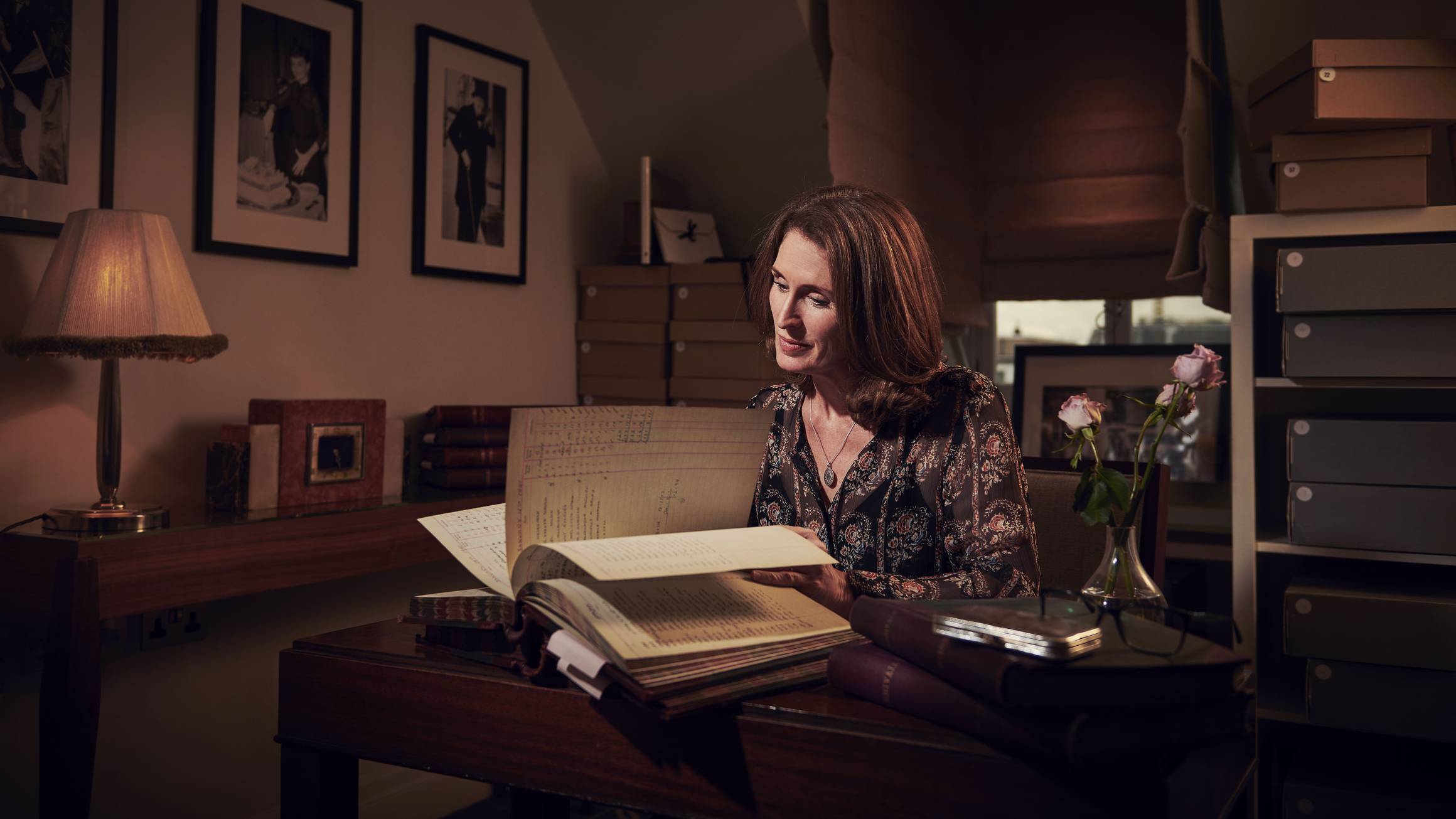 Woman seated at a wooden desk in a softly lit study, reading archival documents, surrounded by books, framed photos, and storage boxes.