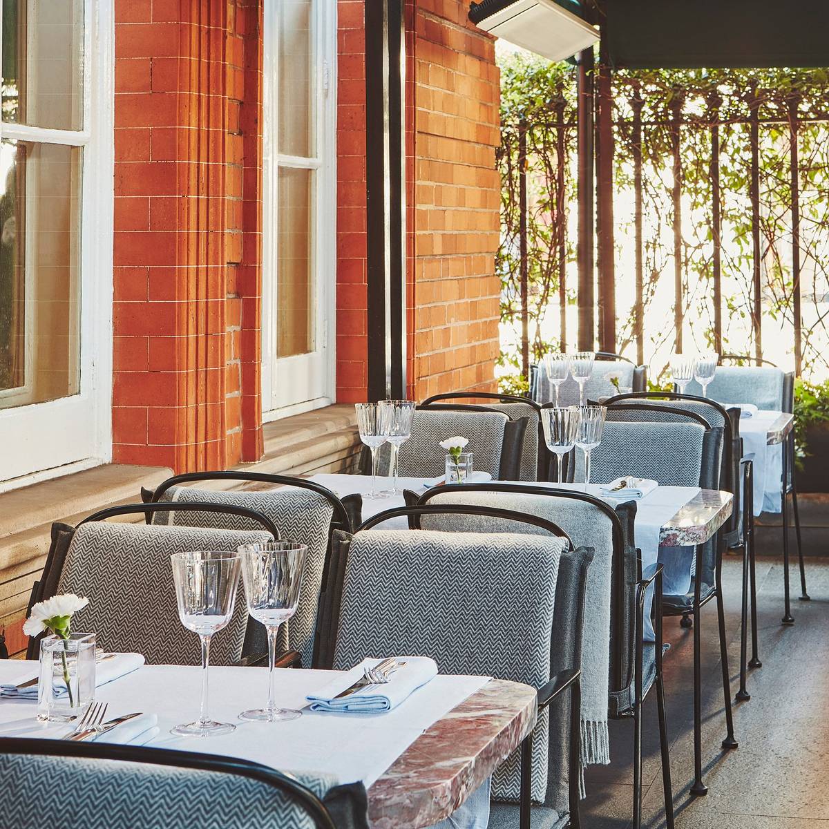 Elegant terrace dining with marble-topped tables, plush chairs draped in blankets, crystal glassware, and lush greenery against red brick walls.
