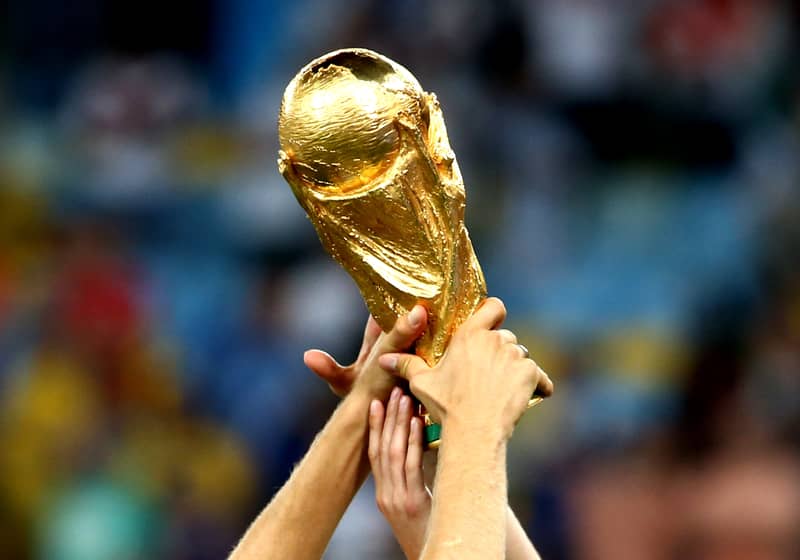Multiple hands raise the golden FIFA World Cup trophy in celebration, with a blurred stadium background.