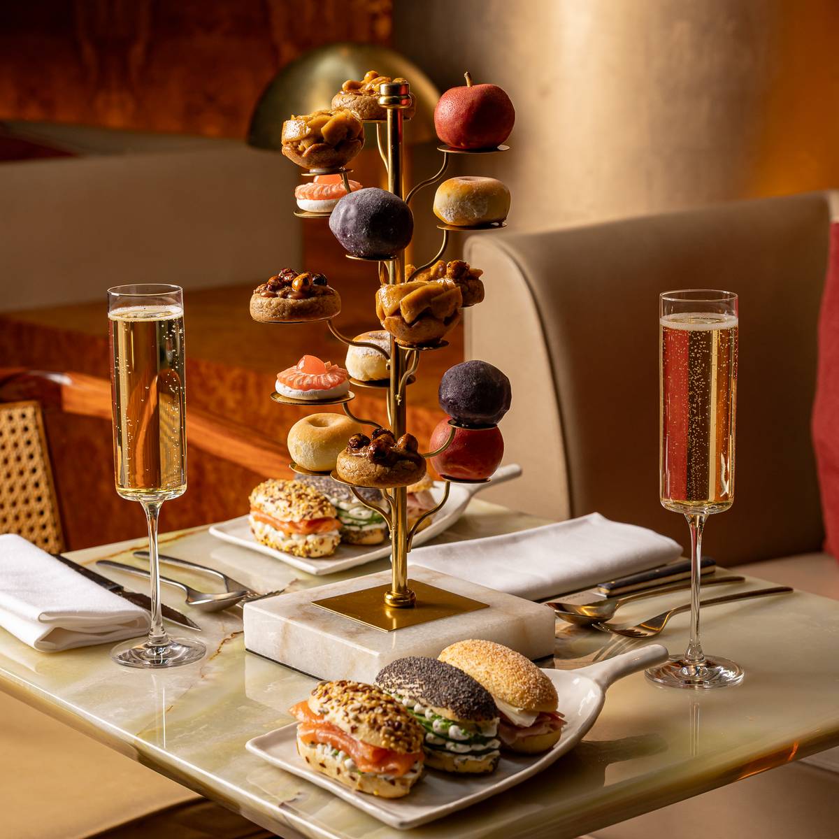 Afternoon tea with fruit-shaped pastries on a golden tree stand, gourmet sandwiches, and two glasses of sparkling wine.
