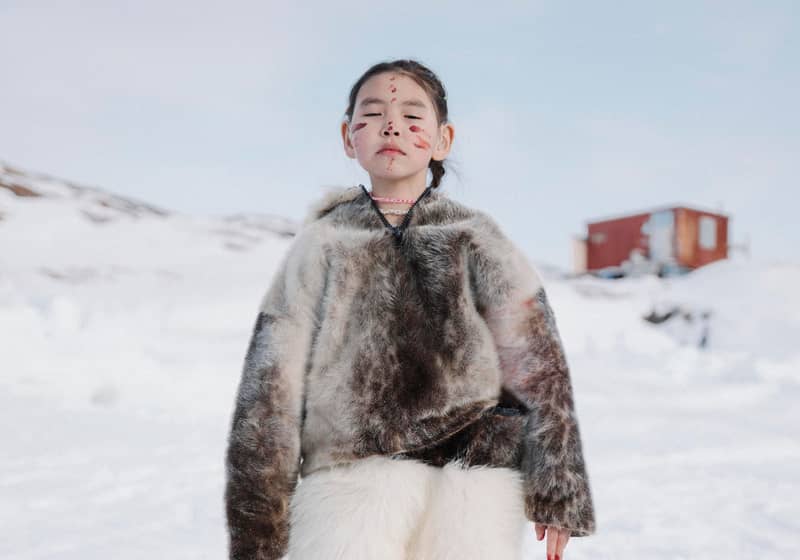 Child in traditional fur clothing stands on snowy ground with face paint, eyes closed, and mountains in background.