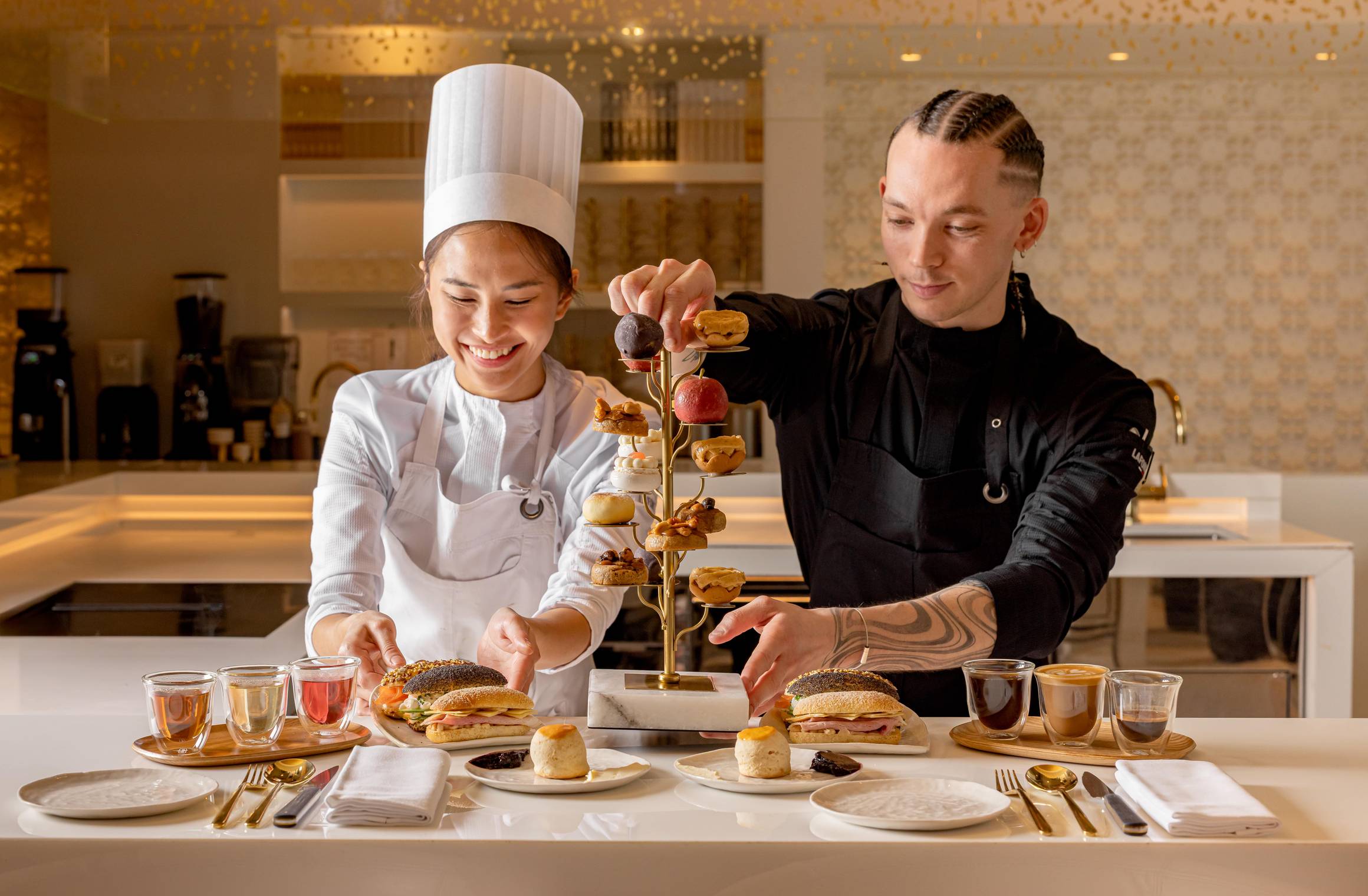 Two smiling chefs at The Berkeley carefully plate an artistic afternoon tea stand with intricate pastries, inside a bright, modern patisserie kitchen.