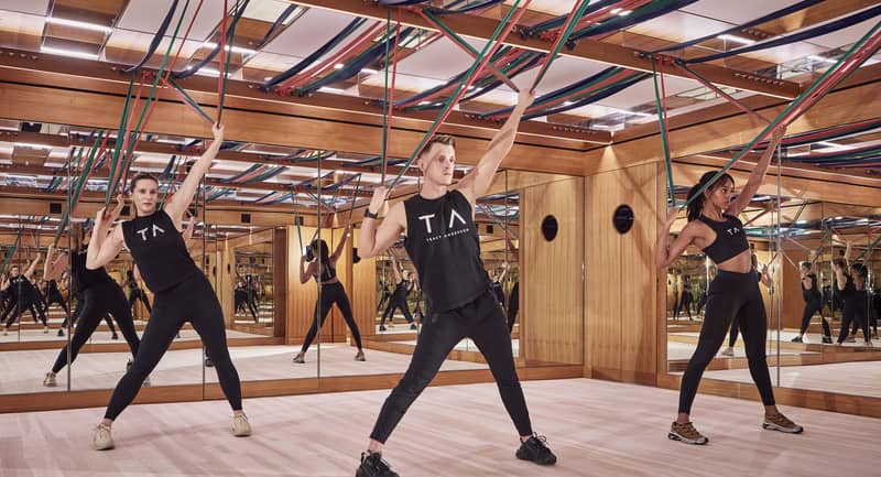 Three people in a luxury studio performing Tracy Anderson Method with resistance bands, surrounded by mirrored walls and wood accents.