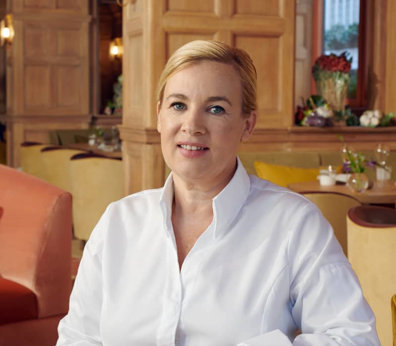 Chef in a white coat seated at a table in an elegant dining room, exuding warmth and professionalism.