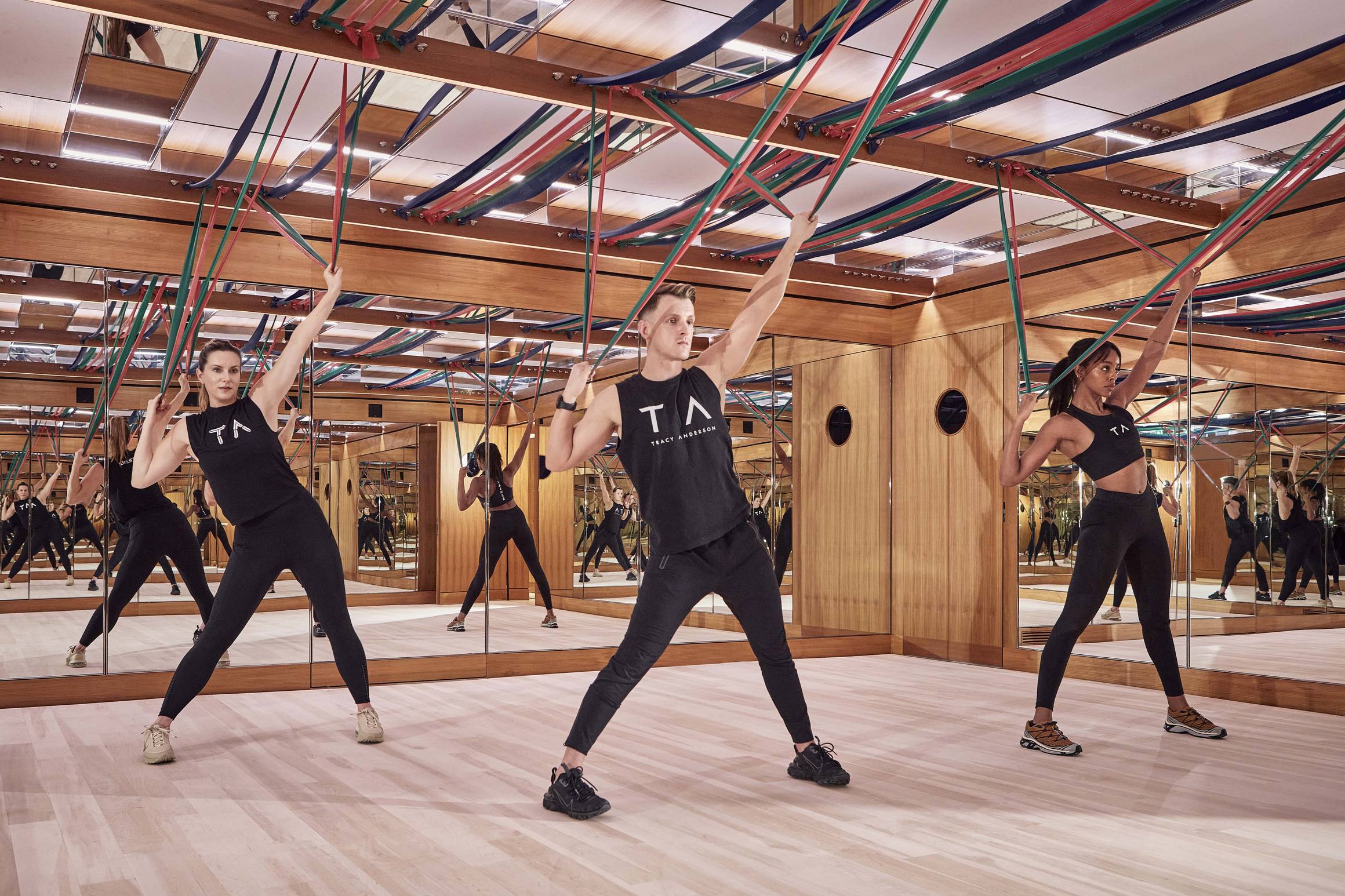 Three people in a luxury studio performing Tracy Anderson Method with resistance bands, surrounded by mirrored walls and wood accents.