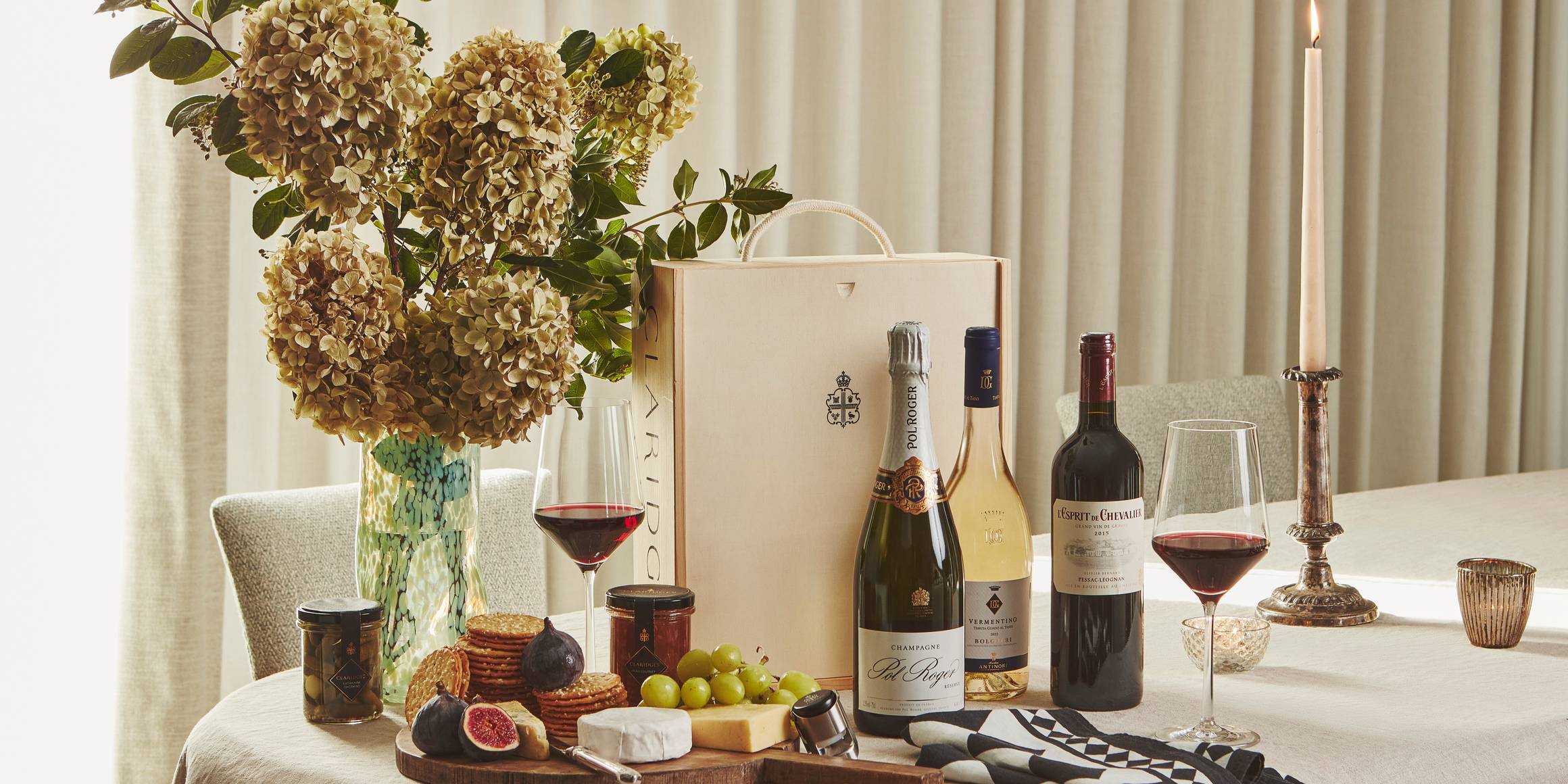 Elegant table with wine, champagne, cheese board, figs, crackers, and flowers, set beside a lit candle and Claridge’s gift box
