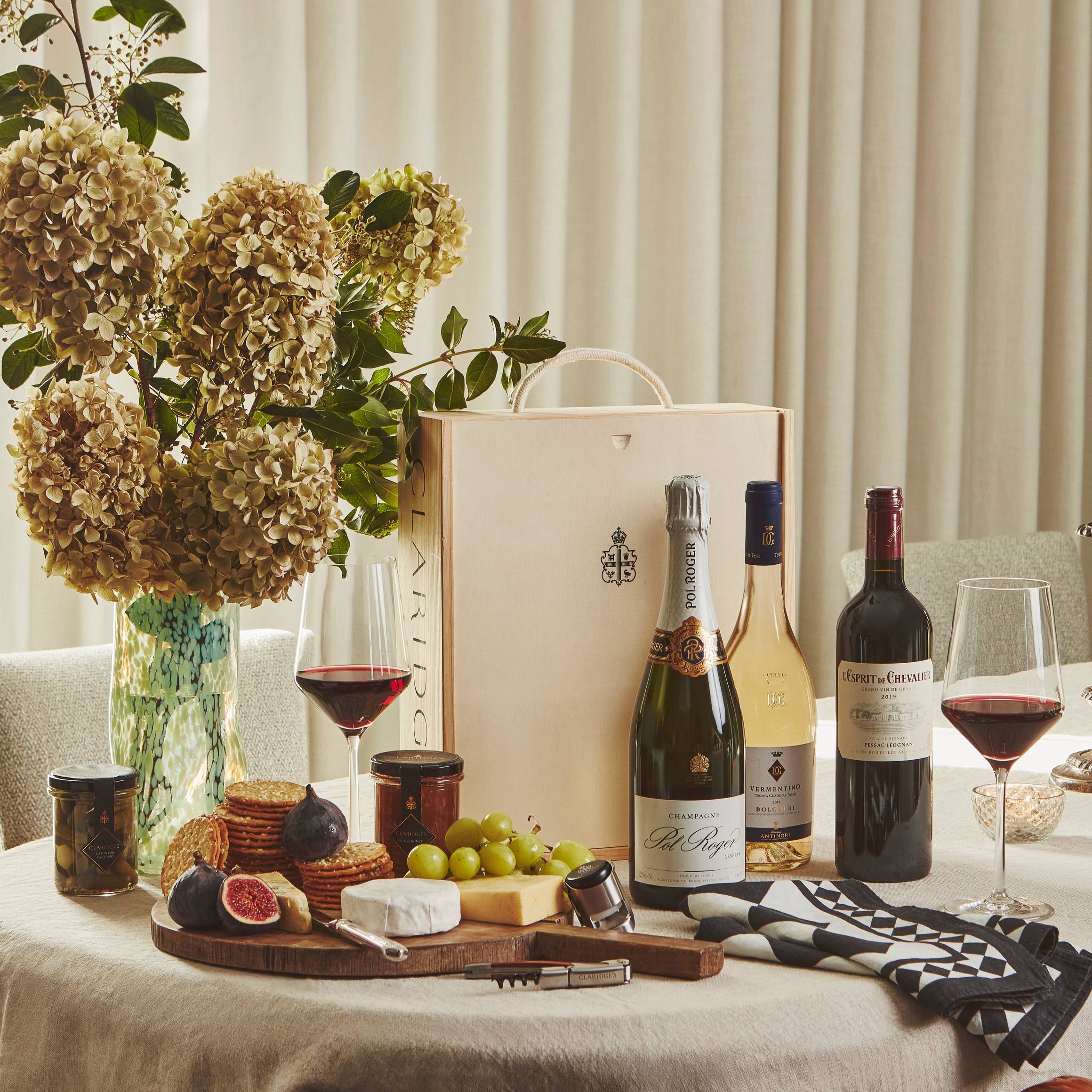 Elegant table with wine, champagne, cheese board, figs, crackers, and flowers, set beside a lit candle and Claridge’s gift box