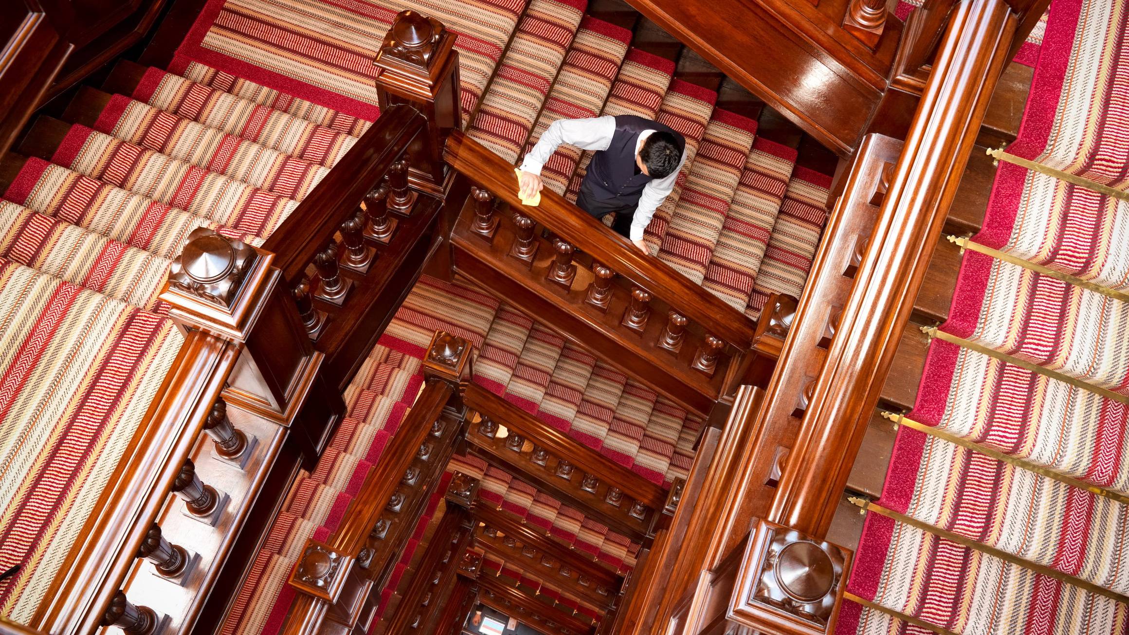 View down The Connaught spiral staircase with polished wood banisters and striped red carpet, as a staff member descends one level mid-frame.