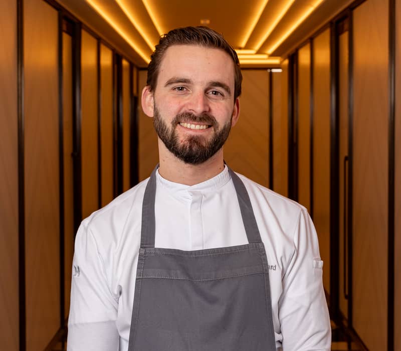 Smiling chef in a white jacket and grey apron stands confidently in a warmly lit, elegant hallway with geometric design
