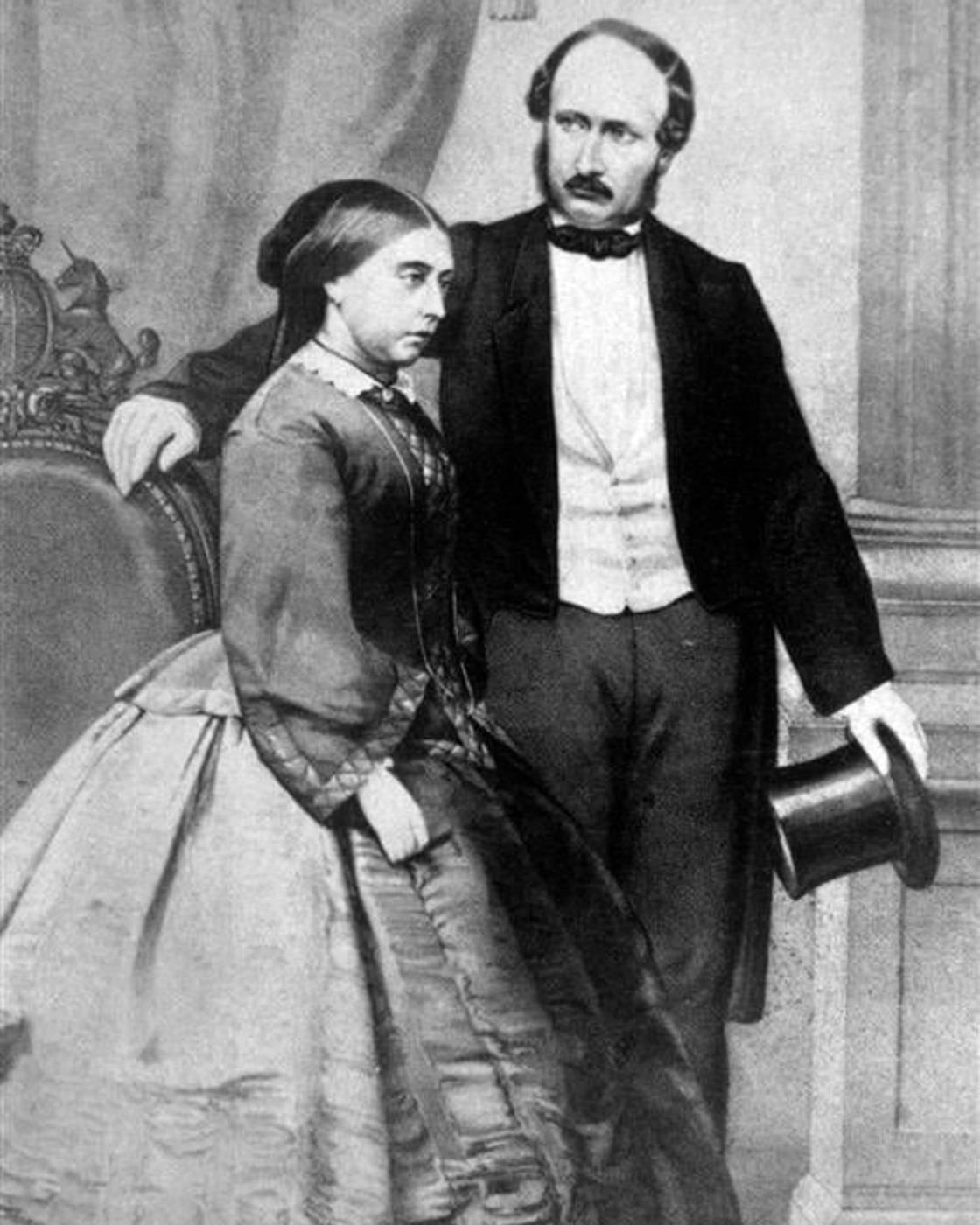 Black and white portrait of a Victorian-era couple; the woman is seated in a full gown, the man stands beside her holding a top hat.