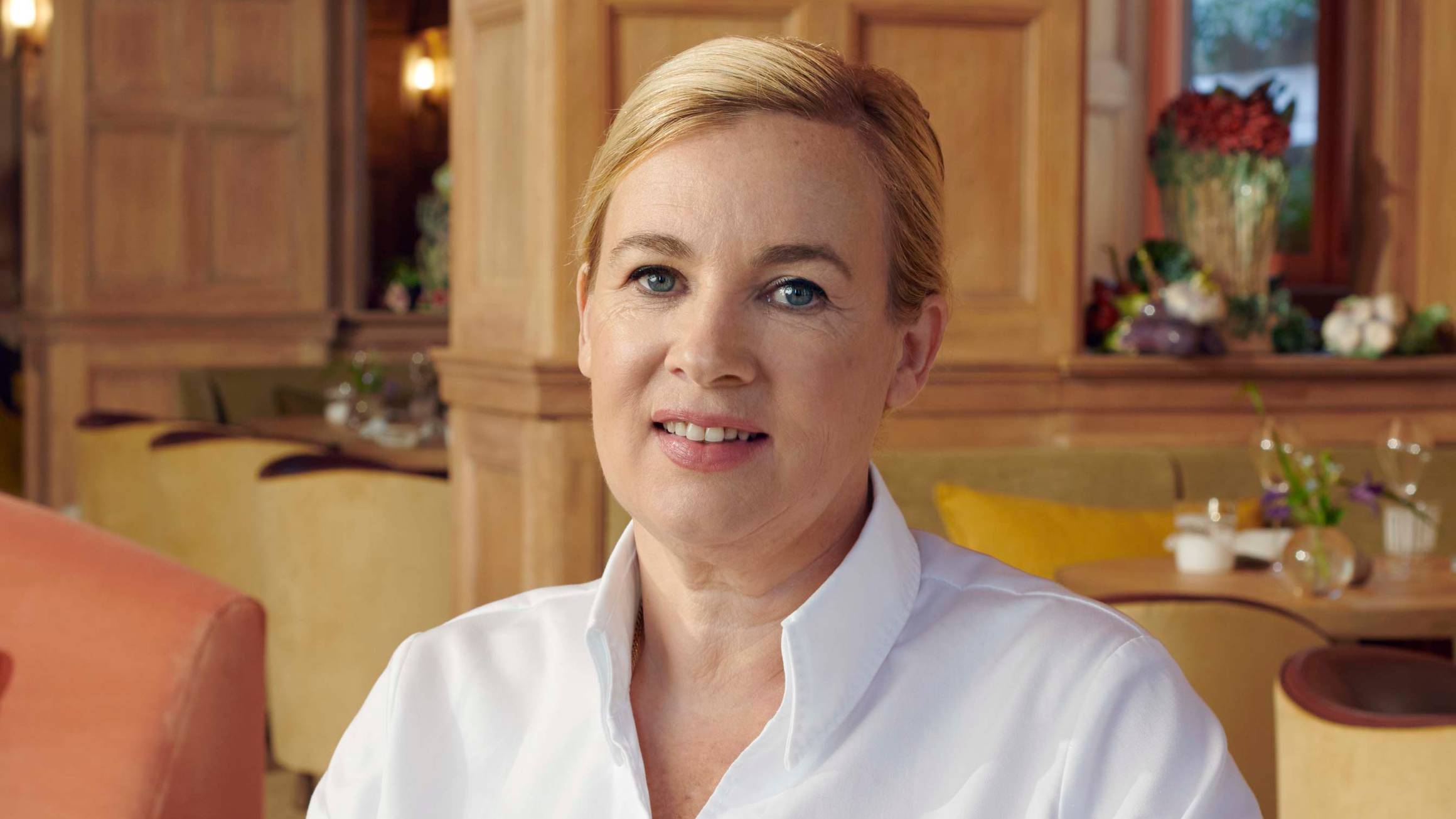 A chef in a white uniform sits at a wooden table in an elegant restaurant with rich wood paneling and soft lighting.