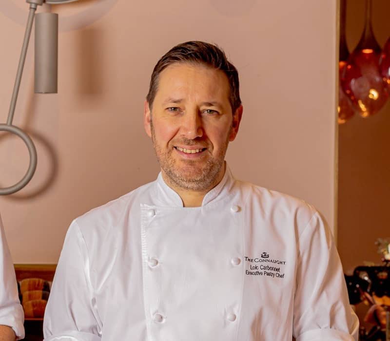 A chef in a white jacket stands proudly behind an elegant pastry display featuring intricate, handcrafted desserts.