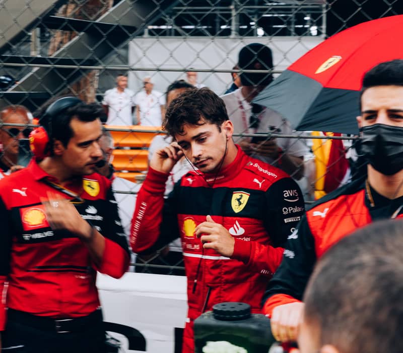 A Formula 1 driver, Charles Leclerc, in a red Ferrari race suit prepares on the grid, surrounded by team members and equipment.