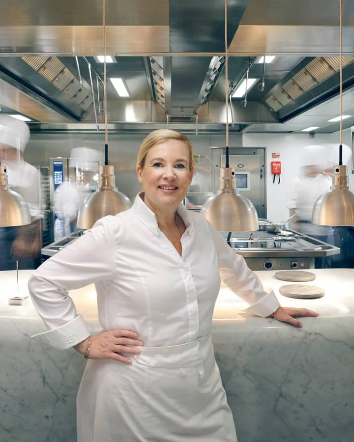A chef in a white uniform stands confidently in a sleek, modern kitchen with stainless steel surfaces and warm lighting.
