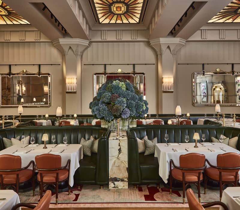 Luxurious restaurant with green leather banquettes, warm lighting, elegant table settings, and a grand floral centerpiece under stained glass.