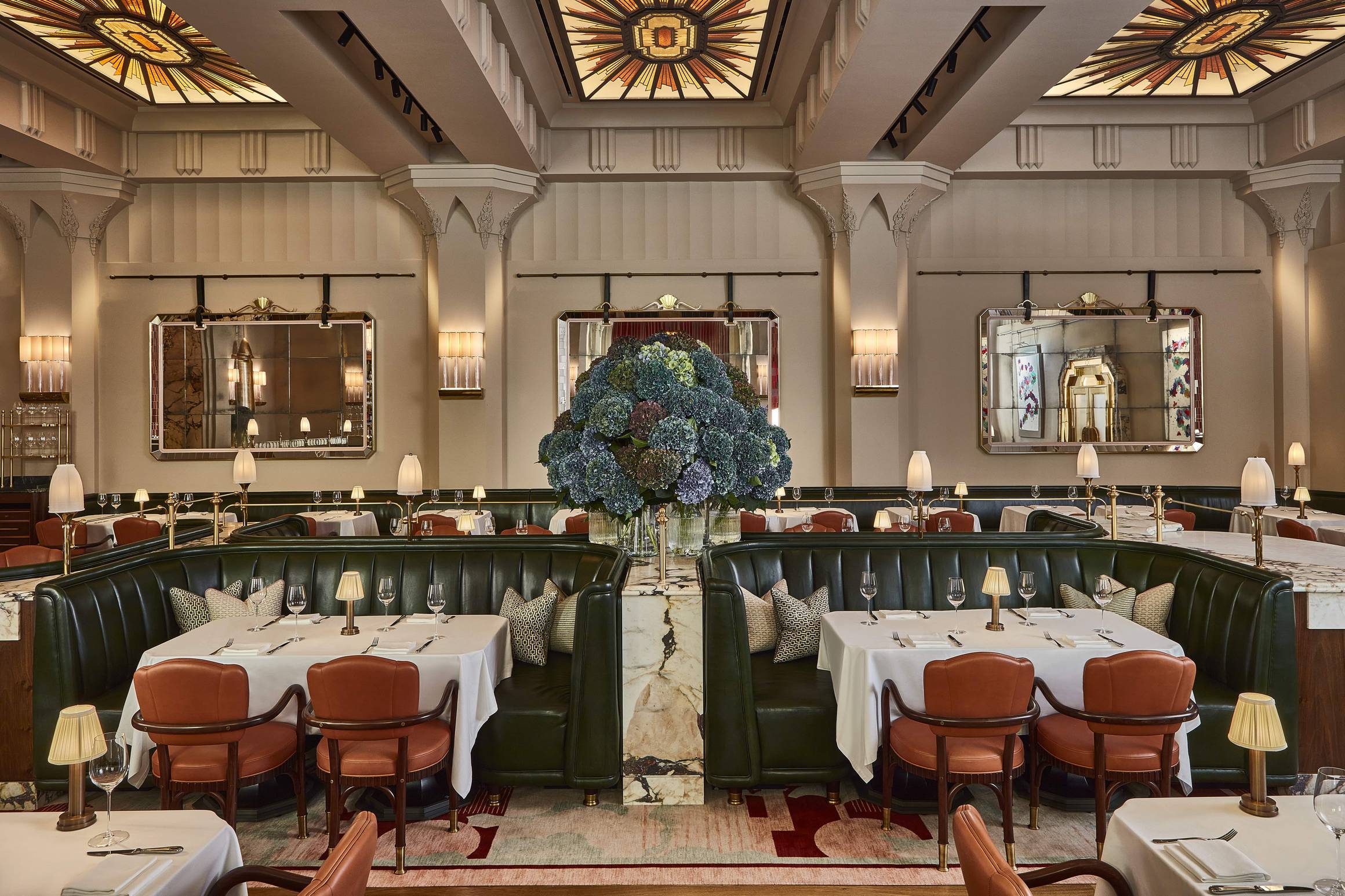 Luxurious restaurant with green leather banquettes, warm lighting, elegant table settings, and a grand floral centerpiece under stained glass.