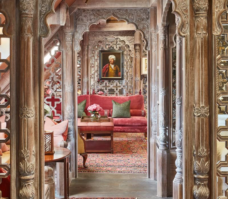 Ornate wooden corridor with carved arches leading to a richly decorated lounge with red sofa, Persian rug, and portrait.