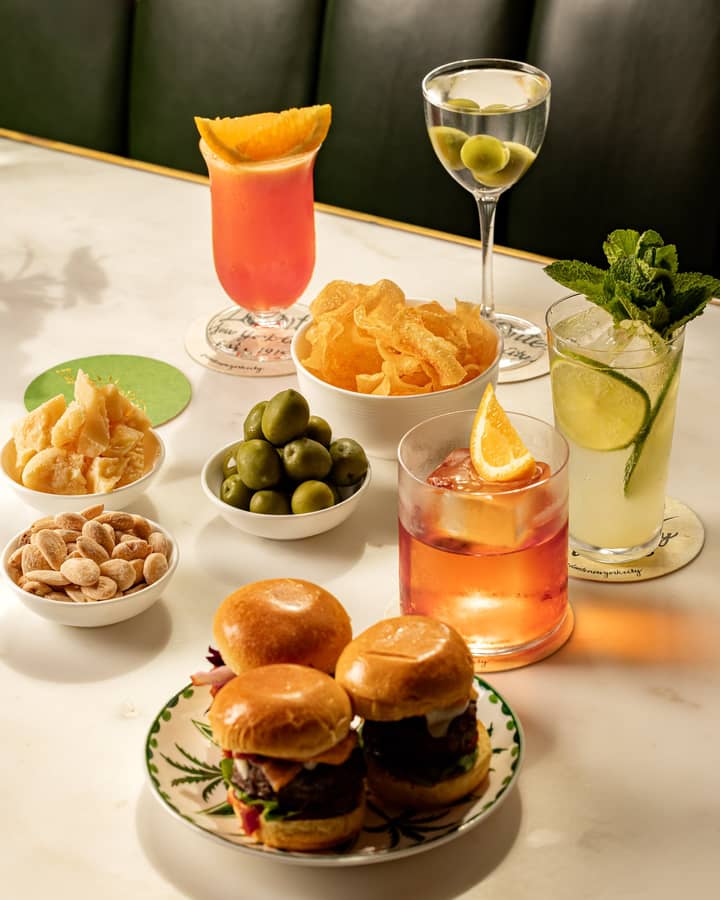 Table with sliders, cocktails, olives, crisps, cheese, and nuts on a white marble surface