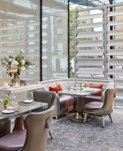 Elegant dining space with leather chairs, marble tables, and a curved banquette set beside large glass windows and floral arrangements.