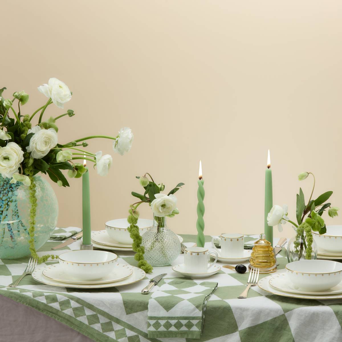 Elegant table setting with white china, green candles, and white ranunculus flowers in vases on a green-and-white patterned cloth.