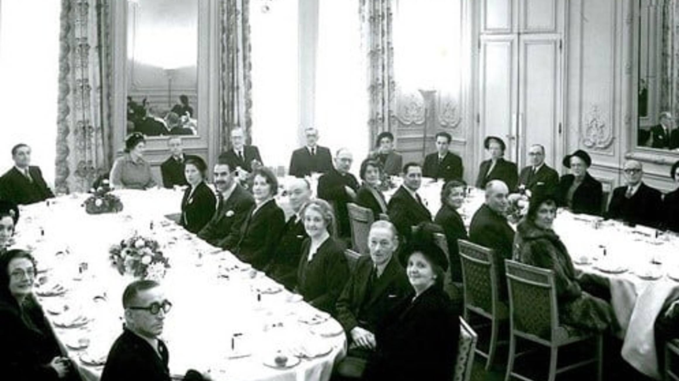 Elegant black-and-white image of guests seated in a grand dining room at Claridge’s, surrounded by chandeliers and ornate decor.