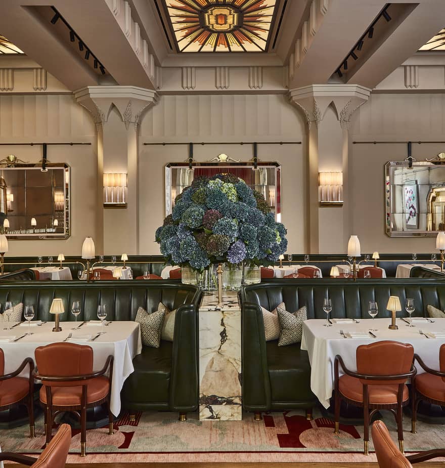 Elegant Art Deco dining room with green leather booths, white tablecloths, and hydrangea centerpiece, under stained glass skylights and symmetrical wall mirrors.