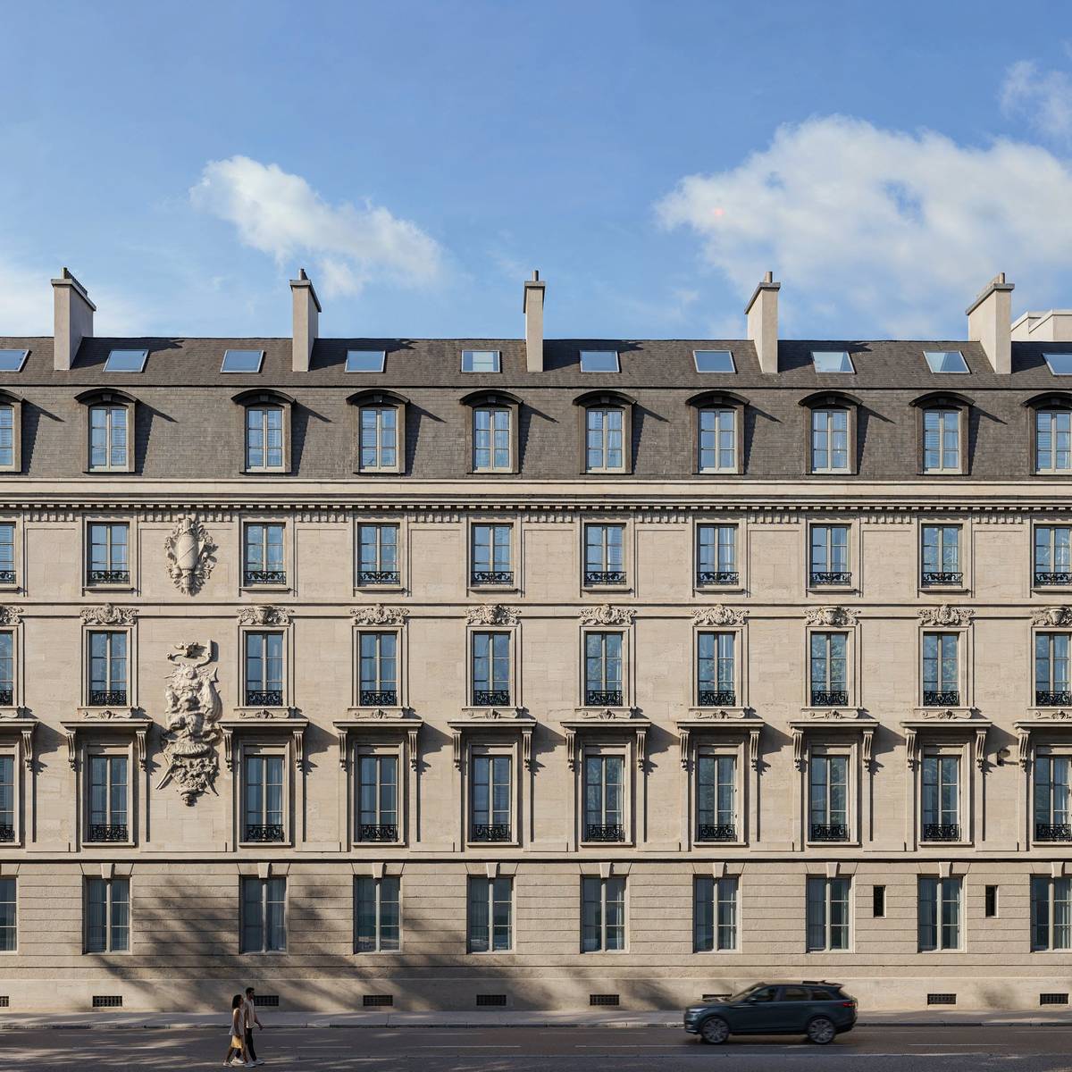 Symmetrical Haussmann-style building with stone carvings, wrought iron balconies, mansard roof, and pedestrians walking along the stree