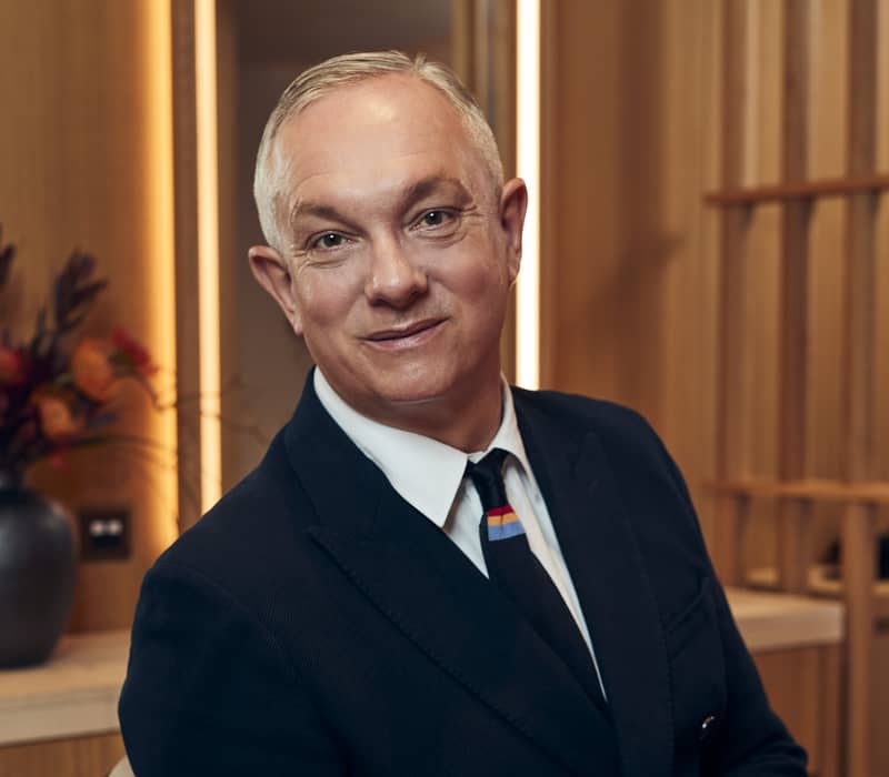 Josh Wood, acclaimed hairstylist, standing confidently in a sleek suit, radiates expertise and elegance in a refined setting.