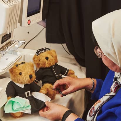 A woman in a blue outfit hand-finishes teddy bears dressed in Claridge’s staff uniforms beside a sewing machine, with one bear wearing a maid outfit and the other in a doorman’s uniform.