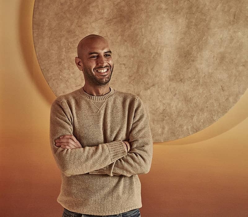 Dr. Mark at Surrenne stands confidently, smiling, in front of a large, textured circle art piece on a warm-toned wall