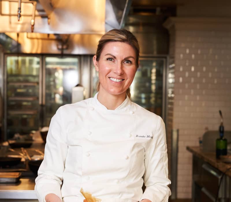 Chef holding a dessert plate with poached pear and ice cream, standing behind a prep counter with ingredients and kitchen tools visible.