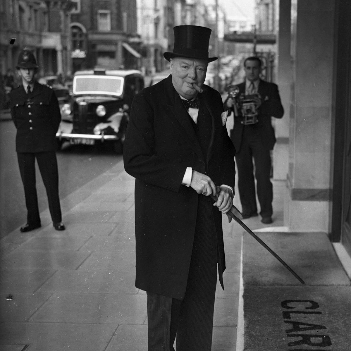 Winston Churchill in top hat and coat with cane and cigar standing outside Claridge’s, vintage car and policeman behind.