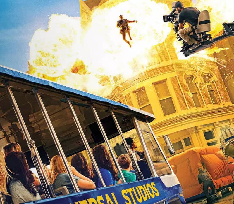 Universal Studios tram ride passes a fiery explosion stunt scene with a cameraman and flying stunt actor.