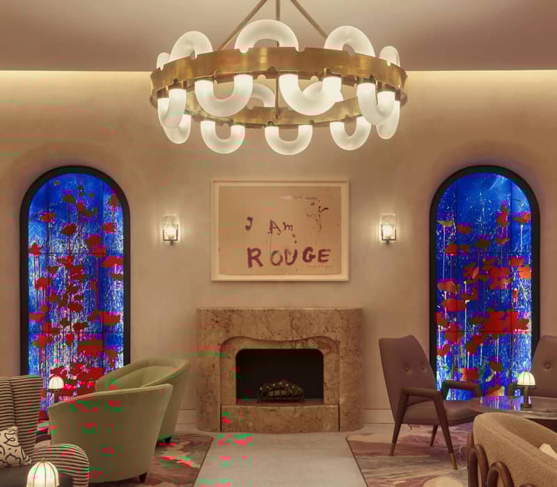 A stylish lounge featuring bold stained glass windows, a marble fireplace, vibrant artwork, and a modern chandelier centerpiece.