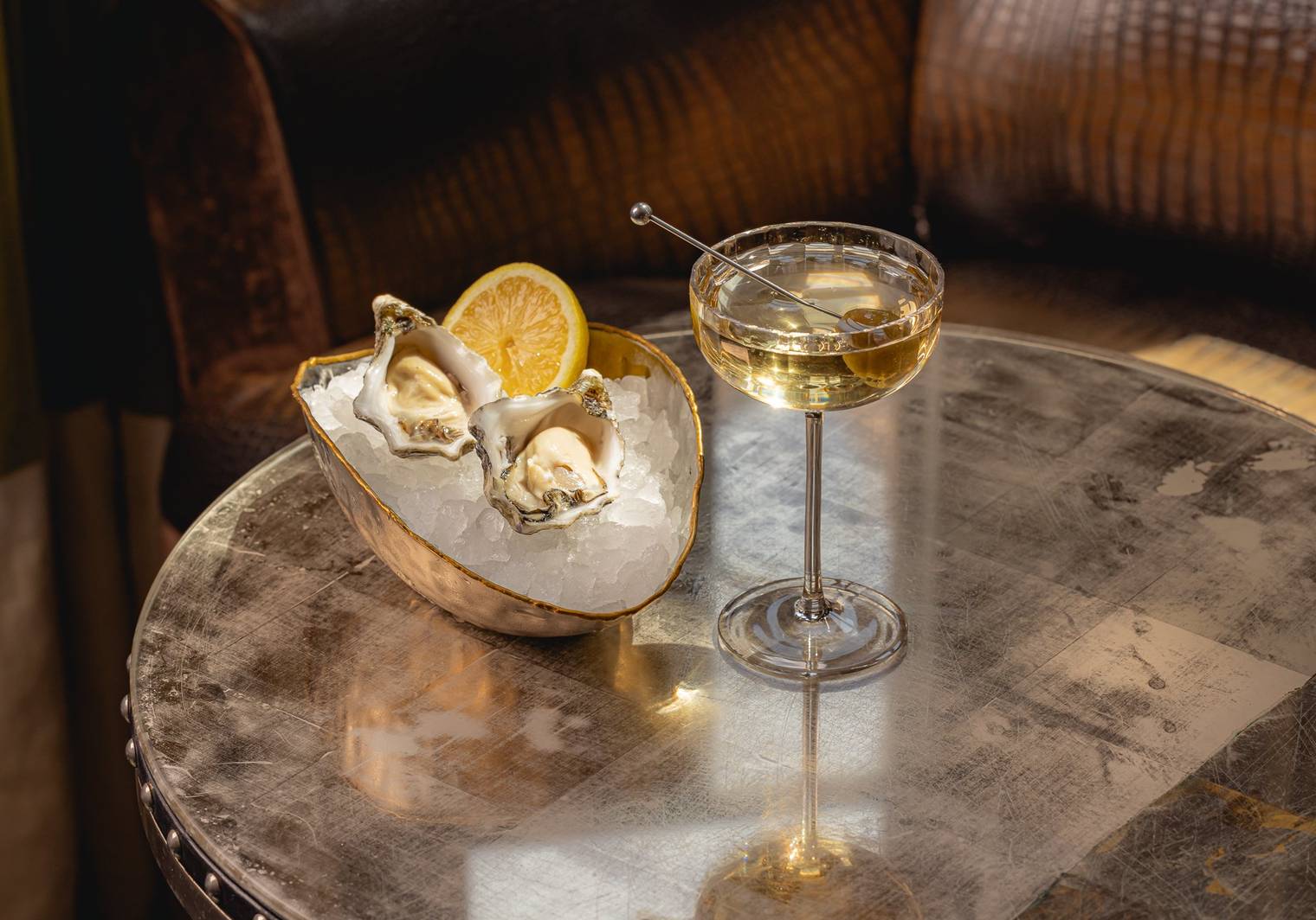 A round table featuring a martini with olives and a gold bowl filled with ice, two oysters and half of a lemon.