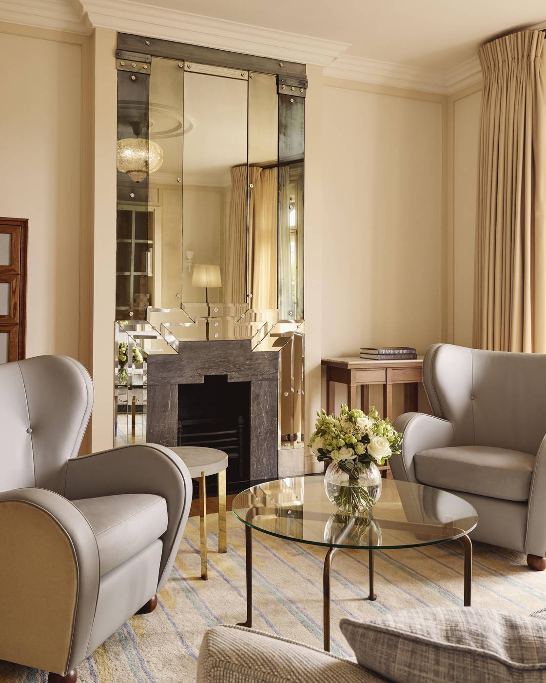 Elegant sitting area in Claridge’s Mayfair Terrace Suite with soft grey armchairs, glass coffee table, Art Deco fireplace, and fresh flowers.